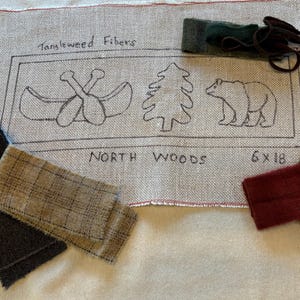May include: A tan fabric with a sketched design of a canoe with paddles, a pine tree, and a bear, with the words "North Woods" and "6 x 18". Several fabric swatches in shades of gray, brown, and burgundy are scattered around the design.