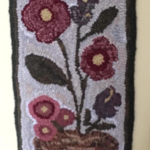 May include: Hand-hooked floral wall hanging featuring a vase with burgundy and pink flowers, dark green leaves, and a purple flower. The background is light gray, and the border is black. The rug is rectangular.