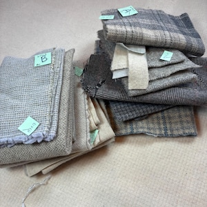 May include: A collection of folded fabric swatches in various shades of gray, beige, and cream. The textiles feature diverse textures including herringbone, waffle weave, and plaid patterns, with small green adhesive labels attached to several pieces.