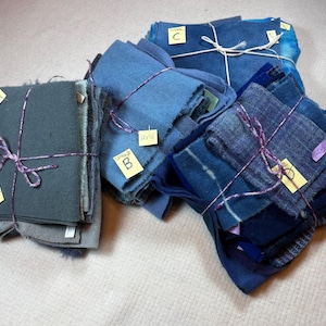 May include: Three bundles of fabric swatches in shades of blue and gray. Each stack is tied with string and features a small yellow paper tag labeled with letters A, B, and C, along with handwritten dimensions in inches.