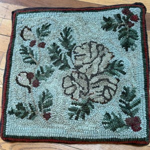 May include: Square, hand-hooked rug with a floral design. The rug features a central flower motif in shades of beige and brown, surrounded by green leaves and red accents. The border is dark brown with a red stripe. The rug is approximately 18 inches square.