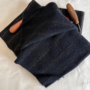 May include: Two pieces of dark fabric, one solid black and the other with a striped pattern, are displayed. Two wooden-handled tools are also present. The fabric appears to be wool or a similar material, and the tools suggest a craft or sewing project.