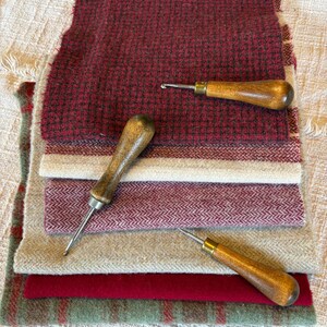 May include: A stack of various textured fabric swatches in shades of red, beige, and green. Three wooden-handled rug hooking tools are placed on top. The fabrics include a red and black check, a herringbone pattern, and solid colors.