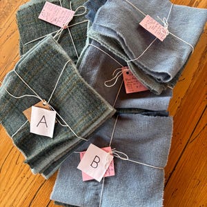 May include: Several bundles of fabric in shades of blue and green are arranged on a wooden surface. Each bundle is tied with white string and has a small tag attached. The fabric appears to be wool or a similar material, with varying textures and patterns.