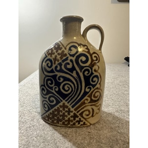 Puede incluir: Una jarra de cerámica beige con una sola asa y cuello estrecho. El cuerpo presenta un patrón decorativo de volutas azul oscuro y diseños geométricos de entramado marrón pintados en la superficie frontal del recipiente.