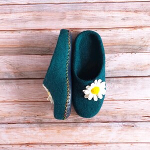 May include: A pair of teal blue felt slippers with white daisy embellishments. The slippers are on a wooden surface.