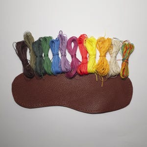 K&ouml;nnte beinhalten: Eine Sammlung von 12 Spulen mit farbigem gewachstem Garn, in Regenbogenform angeordnet, auf einer braunen Lederoberfl&auml;che.