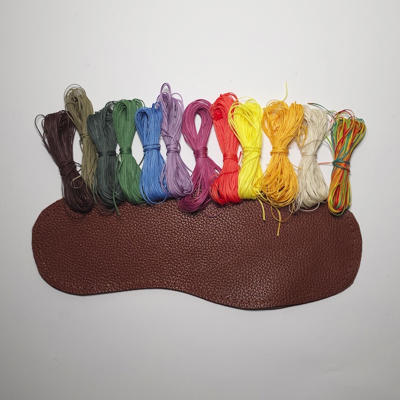 K&ouml;nnte beinhalten: Eine Sammlung von 12 Spulen mit farbigem gewachstem Garn, in Regenbogenform angeordnet, auf einer braunen Lederoberfl&auml;che.