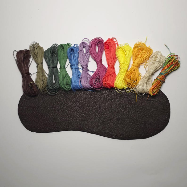 K&ouml;nnte beinhalten: Ein Set mit 12 Spulen gewachsten Fadens in verschiedenen Farben, darunter Rot, Orange, Gelb, Gr&uuml;n, Blau, Lila und Braun. Die Spulen sind in einem Regenbogenmuster auf einer braunen Lederoberfl&auml;che angeordnet.