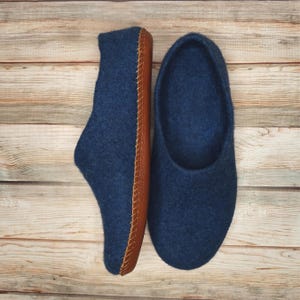 Könnte beinhalten: Ein Paar marineblaue Filzhausschuhe mit braunen Ledersohlen. Einer der Hausschuhe hat eine geschlossene Zehenpartie, der andere ist ein Schlupfschuh. Die Sohlen sind mit einem Kontrastfaden genäht. Der Hintergrund ist helles Holzmaserung.