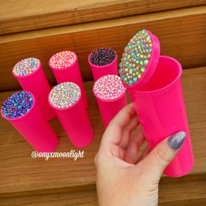 May include: Pink pill containers with clear lids decorated with iridescent rhinestones. The lids are open and the containers are arranged in a row. The text '@onyxmoonlight' is visible in the image.