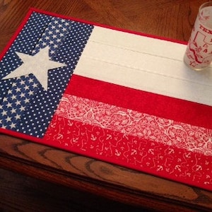 May include: A rectangular fabric placemat featuring a Texas flag design with a blue star section, white stripes, and red stripes with paisley and anchor patterns. A clear glass with red Texas-themed graphics sits on the placemat surface.