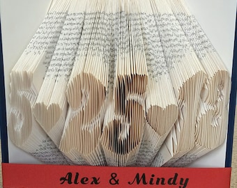 May include: A book art sculpture with the numbers "25" folded into the pages. The book is bound in blue and has a red ribbon with the text "Alex & Mindy" printed on it.