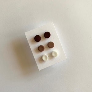 May include: Six small round clay earrings in three colours: brown, light brown, and white. The earrings are arranged in two rows of three on a white card.