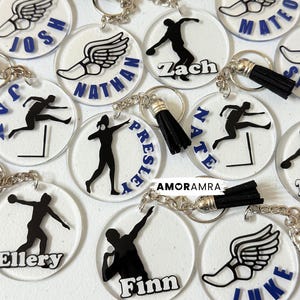 May include: A collection of circular clear acrylic keychains featuring black silhouettes of athletes in track and field poses, including hurdlers and throwers. Each keychain includes a black tassel, a silver metal ring, and a name printed in blue or white.