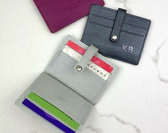May include: Three leather card holders in different colors: purple, gray, and dark blue. The gray card holder is open, showing a credit card and a gift card. The dark blue card holder has the initials "KB" embossed on the front.