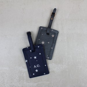 May include: Two luggage tags with a star pattern. One tag is navy blue with white stars and the initials "A.G." printed on it. The other tag is grey with silver stars. Both tags have a black leather strap.