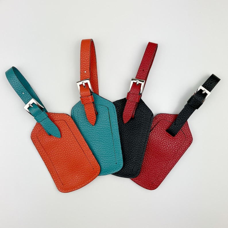 May include: Four leather luggage tags in different colors: orange, teal, black, and red. Each tag has a silver buckle and a loop for attaching to luggage.