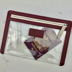 May include: A rectangular clear pouch with a textured burgundy border and a matching zipper closure. The pouch features a small rectangular burgundy tab on the front and contains a burgundy United Kingdom passport with gold lettering and emblem.