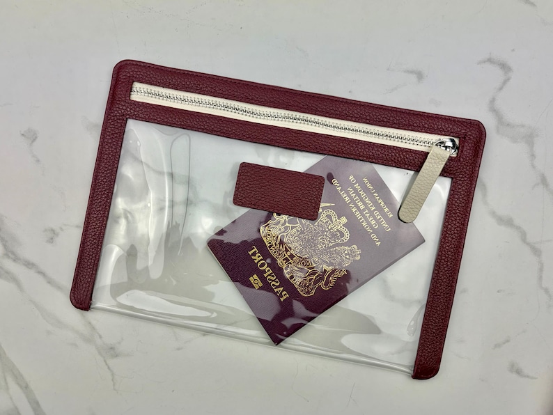 May include: A rectangular clear pouch with a textured burgundy border and a matching zipper closure. The pouch features a small rectangular burgundy tab on the front and contains a burgundy United Kingdom passport with gold lettering and emblem.