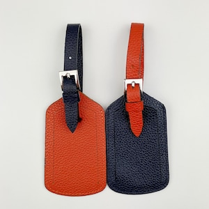 May include: Two leather luggage tags, one orange and one navy blue. Both tags have a silver metal loop and a silver metal buckle.