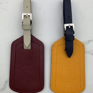 May include: Two leather luggage tags with a burgundy and a yellow tag. The burgundy tag has a white strap and the yellow tag has a navy blue strap. Both tags have a silver buckle.