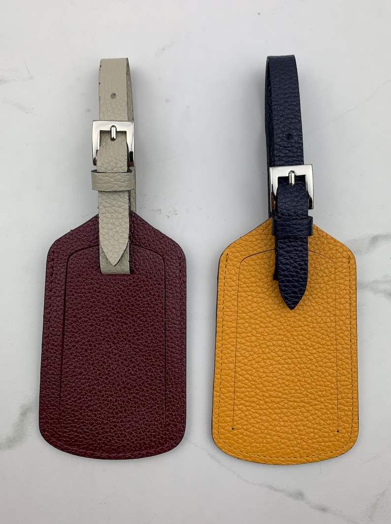 May include: Two leather luggage tags with a burgundy and a yellow tag. The burgundy tag has a white strap and the yellow tag has a navy blue strap. Both tags have a silver buckle.