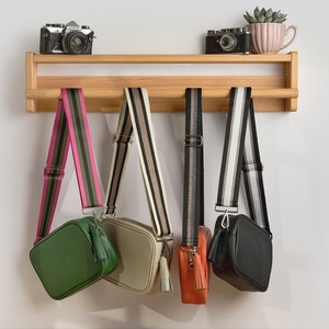 Puede incluir: Tres bolsos de bandolera de diferentes colores con correas a rayas colgando de una estantería de madera. Los bolsos son verde, beige y negro. La estantería tiene dos cámaras y una pequeña planta en maceta.