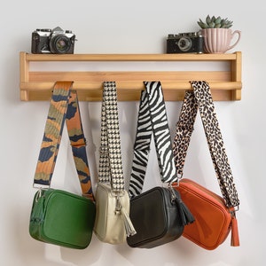 May include: Five different patterned crossbody bags hanging on a wooden shelf with two vintage cameras and a potted succulent on the top shelf.
