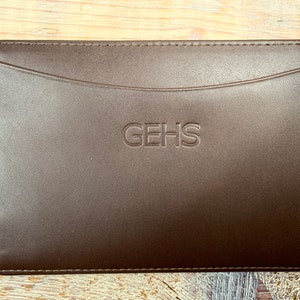 Puede incluir: Una bolsa de cuero marr&oacute;n con un borde cosido y un solo bolsillo. La bolsa tiene la palabra "GEHS" grabada en la parte delantera.