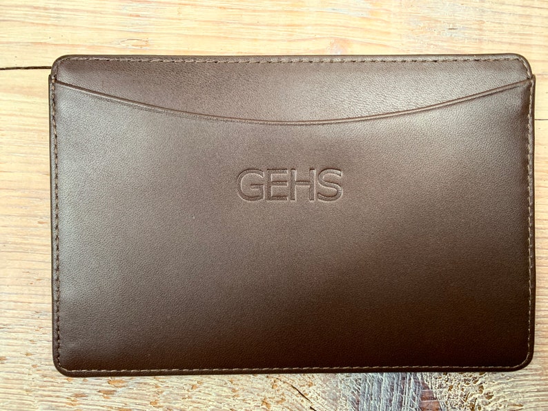 Puede incluir: Una bolsa de cuero marr&oacute;n con un borde cosido y un solo bolsillo. La bolsa tiene la palabra "GEHS" grabada en la parte delantera.