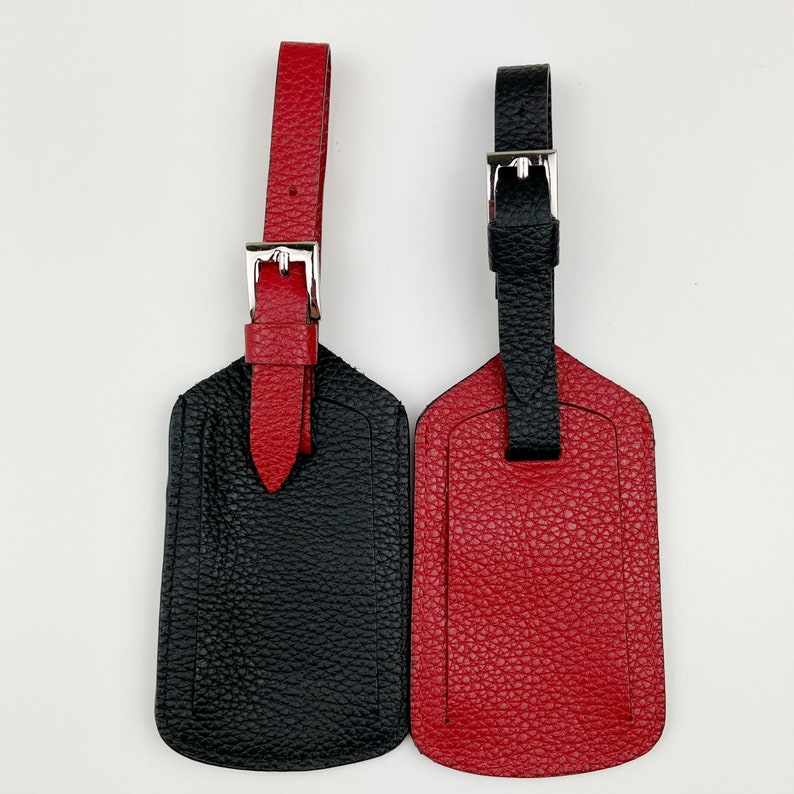 May include: Two leather luggage tags, one black and one red, with silver buckles. The tags are made of textured leather and have a simple design.