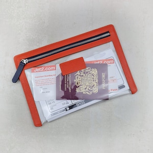May include: A rectangular clear travel pouch with a textured orange leather border and a dark blue zipper. Inside the pouch are Jet2.com boarding passes and a burgundy United Kingdom passport with the royal coat of arms visible on the cover.