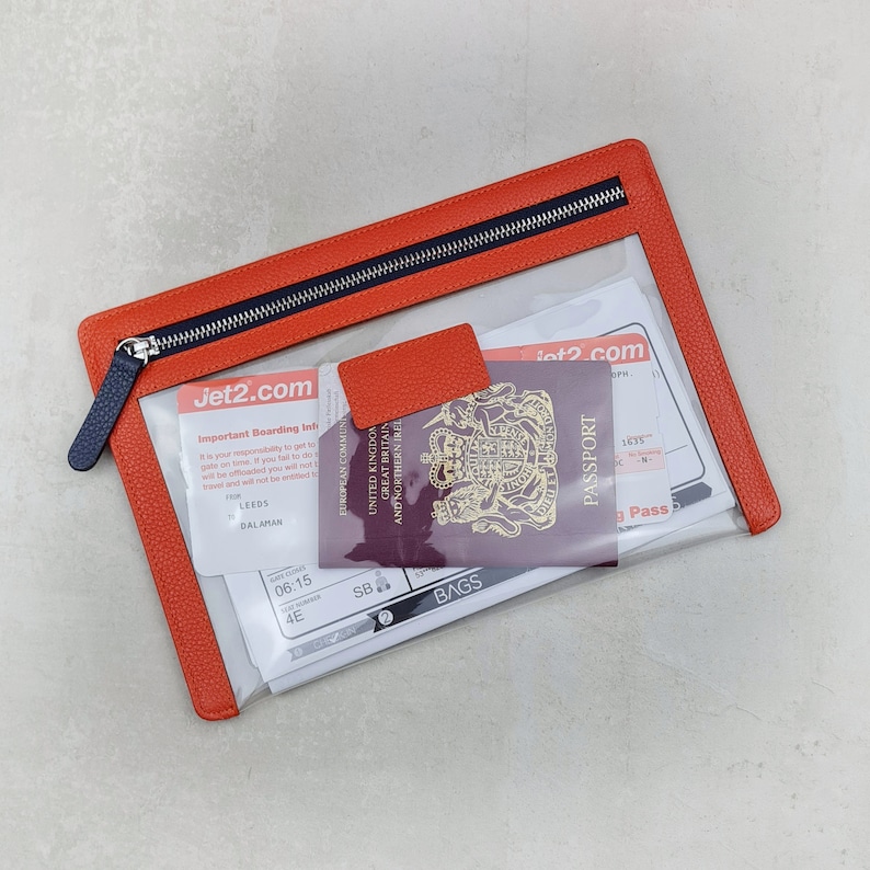 May include: A rectangular clear travel pouch with a textured orange leather border and a dark blue zipper. Inside the pouch are Jet2.com boarding passes and a burgundy United Kingdom passport with the royal coat of arms visible on the cover.
