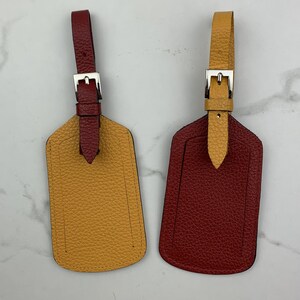 May include: Two leather luggage tags with metal buckles. One tag is mustard yellow and the other is burgundy red.