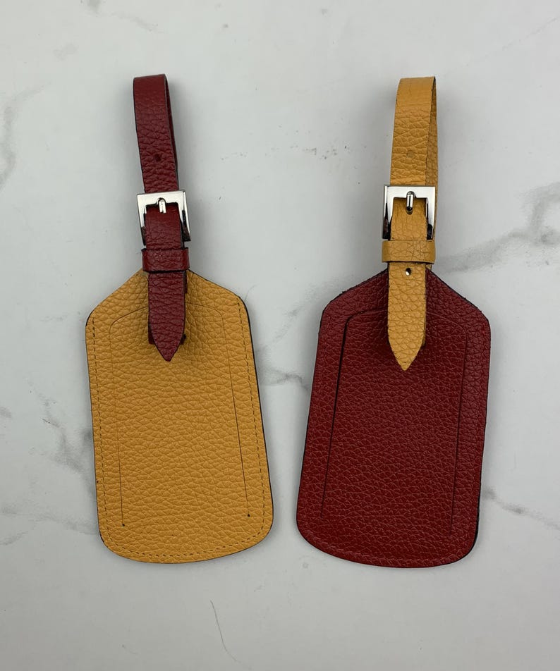May include: Two leather luggage tags with metal buckles. One tag is mustard yellow and the other is burgundy red.