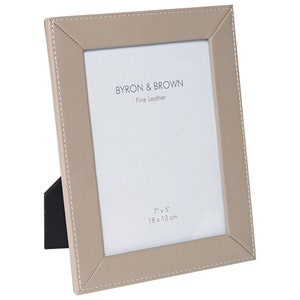 May include: A beige leather picture frame with a stitched border. The frame is 7 inches by 5 inches, or 18 cm by 13 cm. The frame has a black stand on the back. The frame says "BYRON & BROWN Fine Leather" on the front.