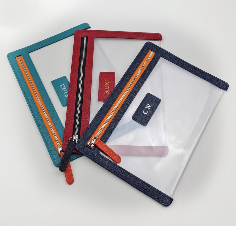 May include: Three rectangular clear pouches with textured leather-like borders in teal, red, and navy blue. Each pouch features a metal zipper with a matching pull tab and a small rectangular tag. The tags are embossed with the text RUKI or CW.