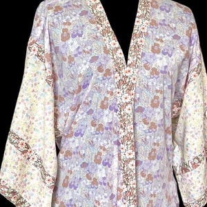 May include: A floral print kimono in shades of lavender, purple, and brown. The kimono features a V-neck and wide sleeves. The edges are trimmed with a floral pattern in white and pink.