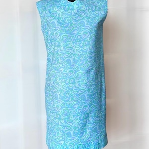May include: A sleeveless shift dress featuring a repeating abstract pattern in shades of light blue, turquoise, and lavender. The garment has a high, rounded neckline and a straight, knee-length silhouette, displayed on a black mannequin torso.