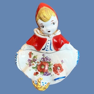 May include: A vintage ceramic cookie jar shaped like a girl wearing a red hooded cloak. The jar has a white base with a floral design and a yellow basket.