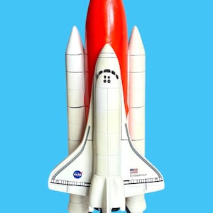 May include: A model of the NASA Space Shuttle Endeavour featuring a white orbiter with black cockpit windows, a large orange external fuel tank, and two white solid rocket boosters. The shuttle displays the NASA logo and the name Endeavour.