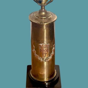 May include: A brass table lamp with a cylindrical base and a black square base. The lamp features a decorative emblem with a crest and laurel leaves. The lamp has a gold finish and a standard lamp shade fitting.