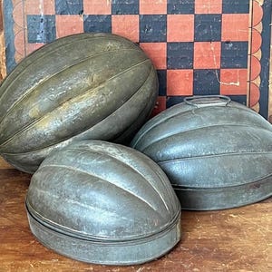 May include: Three antique metal melon molds. The molds are oval-shaped with ribbed detailing and a weathered, aged appearance. One mold has a handle on top. The background includes a vintage checkerboard.