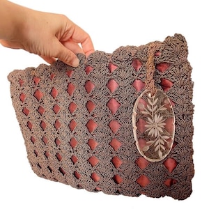 Könnte beinhalten: Eine Vintage-Handtasche aus braunem Häkelwerk mit einem rosafarbenen Satinfutter. Die Tasche hat einen dekorativen ovalen Anhänger aus klarem Glas mit einem geätzten Blumendesign. Die Tasche wird von einer Hand gehalten.