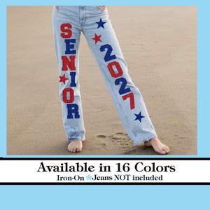 Op de afbeelding: Lichtblauwe spijkerbroek met rode en blauwe glitterletters met de tekst "SENIOR 2027" en steraccenten. De spijkerbroek wordt getoond op een persoon die op een zandstrand staat. De tekst "Available in 16 Colors" staat onderaan.