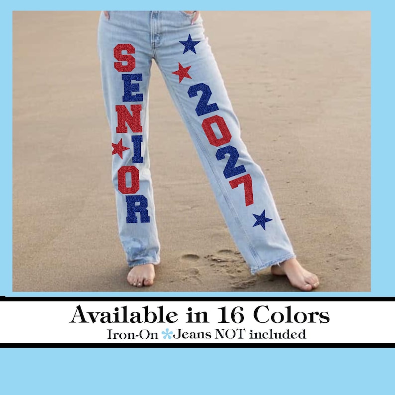 Op de afbeelding: Lichtblauwe spijkerbroek met rode en blauwe glitterletters met de tekst "SENIOR 2027" en steraccenten. De spijkerbroek wordt getoond op een persoon die op een zandstrand staat. De tekst "Available in 16 Colors" staat onderaan.