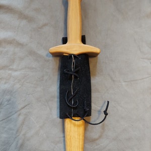 May include: A wooden dagger with a black leather sheath and a knotted strap. The dagger has a rounded handle and a pointed blade.