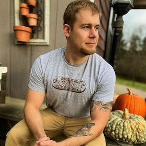 Puede incluir: Un hombre con una camiseta gris con un logotipo marrón y blanco que dice "BORDERLINE" y "CITY OF DETROIT". Está sentado en un escalón de madera con una calabaza y una calabaza a su lado.