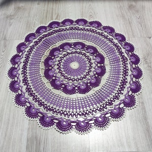 Op de afbeelding: Een groot, rond, gehaakt tafellaken met een paars en wit ontwerp. Het tafellaken heeft een centrale cirkel met meerdere lagen van ingewikkelde gehaakte patronen.